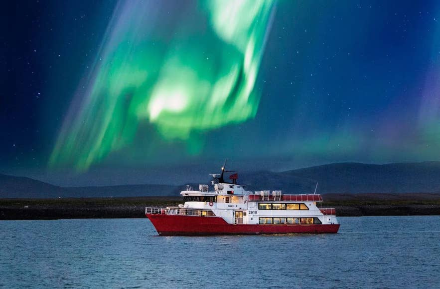 Green northern lights above a red and white boat sailing on the ocean off the coast of Iceland
