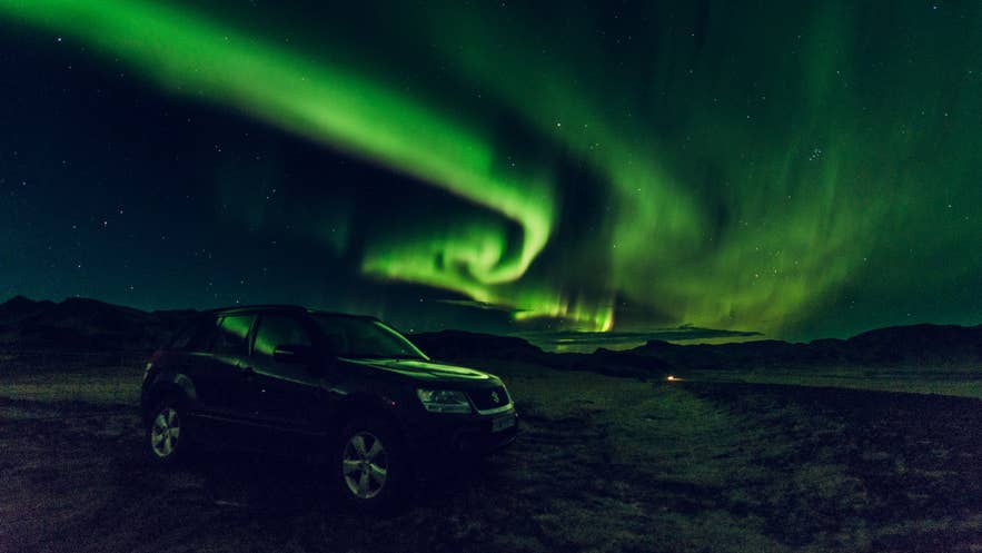 A parked SUV under a swirling green aurora in the Icelandic countryside at night