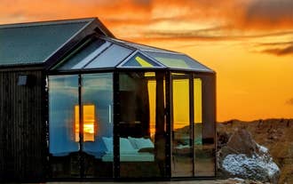 Glass cabin lit from within under an orange sunset sky on a farm in South Iceland.