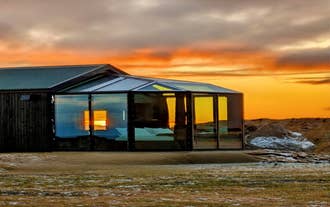 Glass cabin lit from within under an orange sunset sky on a farm in South Iceland.