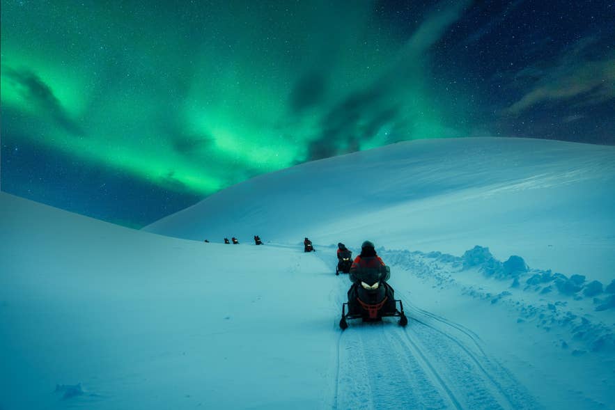 A line of travelers on snowmobiles crossing a snow covered glacier under the northern lights in Iceland.