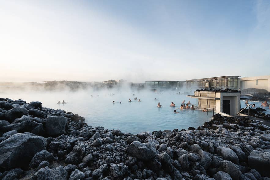 Lava rocks frame the steaming water of Blue Lagoon Geothermal Spa as bathers soak beside the spa buildings.
