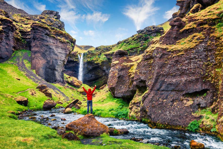 A hiker in a red jacket on the riverside trail leading to Kvernufoss waterfall in summer.