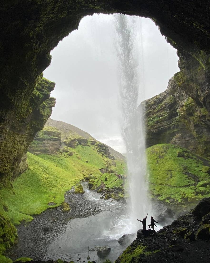 Two visitors standing behind the curtain of water at Kvernufoss waterfall in summer, seen from inside the cave.
