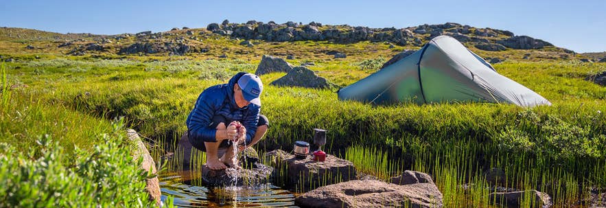 A hiker using camping gear during an overnight stop in the Icelandic countryside, with a tent and cookware set up beside a stream.