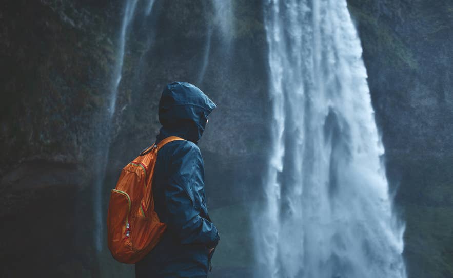 A traveler in a blue hooded waterproof jacket and orange backpack stands in the spray of a waterfall. A traveler in a blue hooded waterproof jacket and orange backpack stands in the spray of a waterfall.