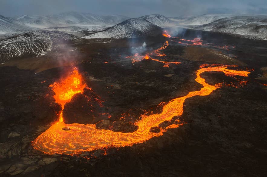 A river of molten lava flowing from an erupting vent across dark ground with snowy mountains behind