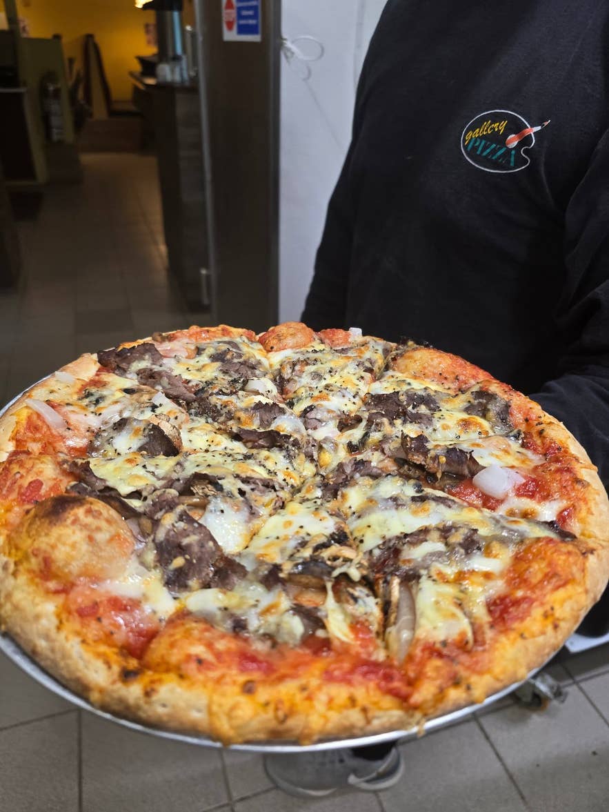 Staff member holds a freshly baked pizza topped with meat and mushrooms at Gallery Pizza in Hvolsvollur.