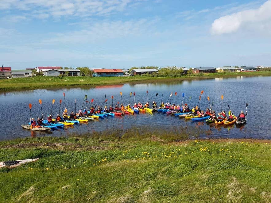 A large group of kayakers lined up on a calm lake beside a village in Iceland A large group of kayakers lined up on a calm lake beside a village in Iceland