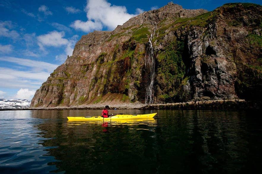 A kayaker paddles beneath a steep cliff with a waterfall in Iceland A kayaker paddles beneath a steep cliff with a waterfall in Iceland