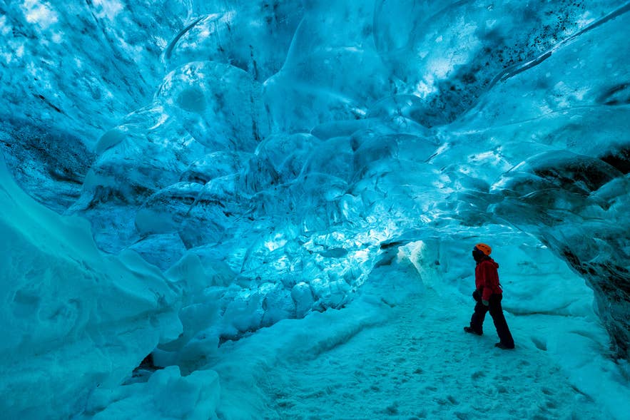 Person in a red jacket and orange helmet walking through a blue ice cave