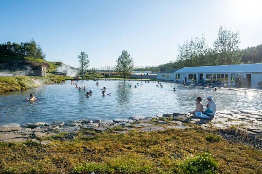 Bathers in the Secret Lagoon on a sunny day, with the changing building and trees behind the pool