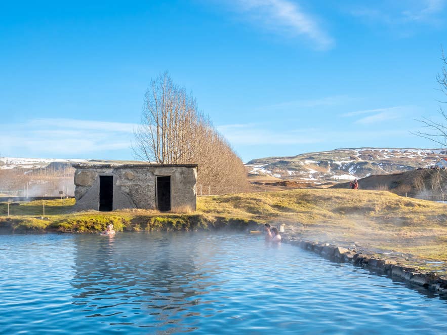 The old stone building at the Secret Lagoon under blue sky, with steam over the pool and snowy hills behind
