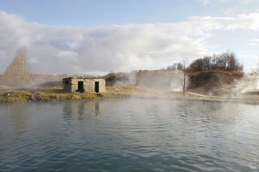 Steam drifting across the Secret Lagoon toward the old stone building on a bright day