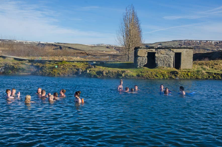 A group of bathers in the Secret Lagoon on a sunny day, with the old stone building and a tall tree behind