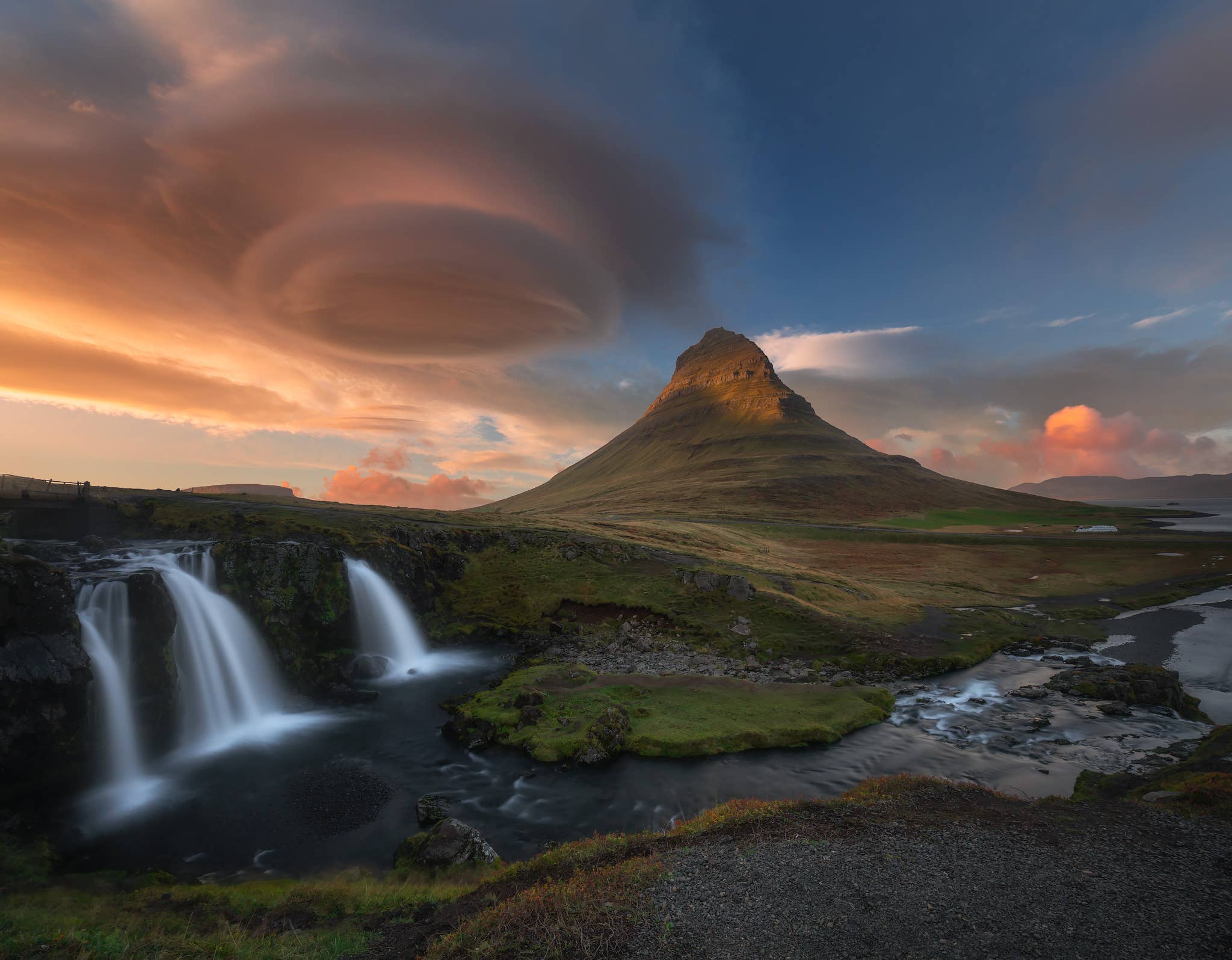 Kirkjufell Mountain rises behind Kirkjufellsfoss Waterfall under a lenticular cloud at sunset on the Snaefellsnes Peninsula.