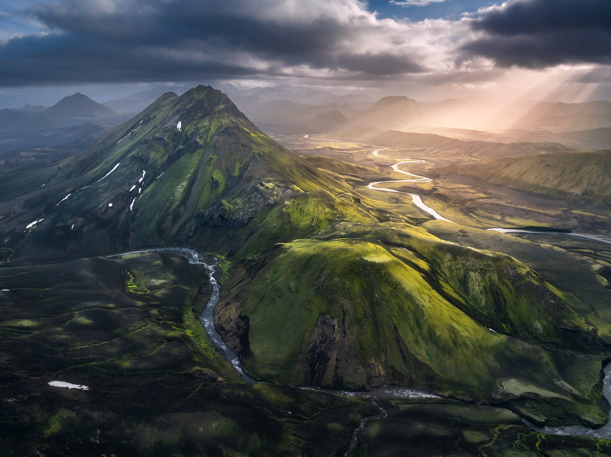 Sun rays break through clouds over green mountains and a winding river near the Laugavegur Trail in the Highlands.