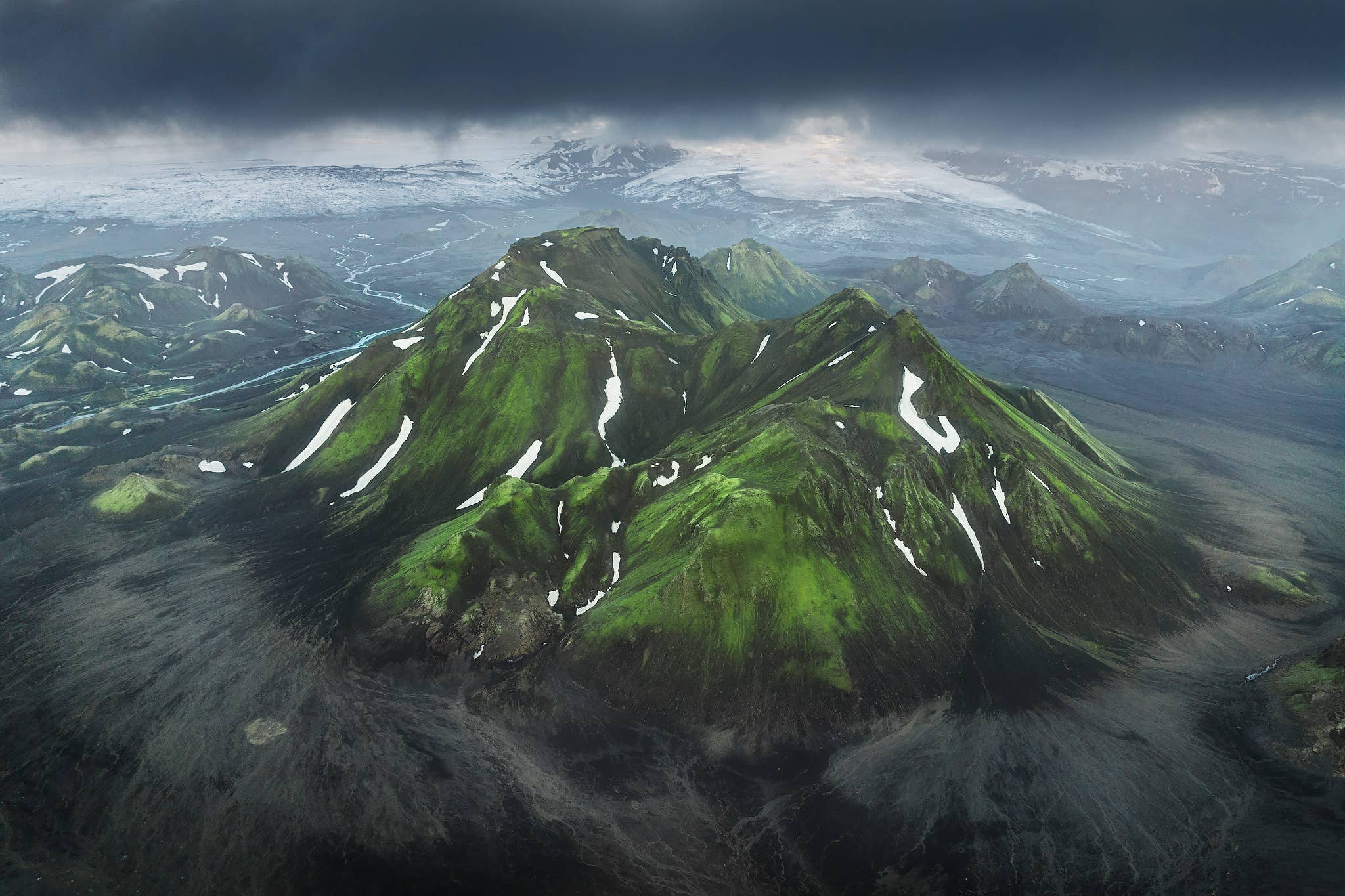 Snow-streaked, moss-covered mountains rise below dark clouds near the Laugavegur Trail in the Highlands of Iceland.