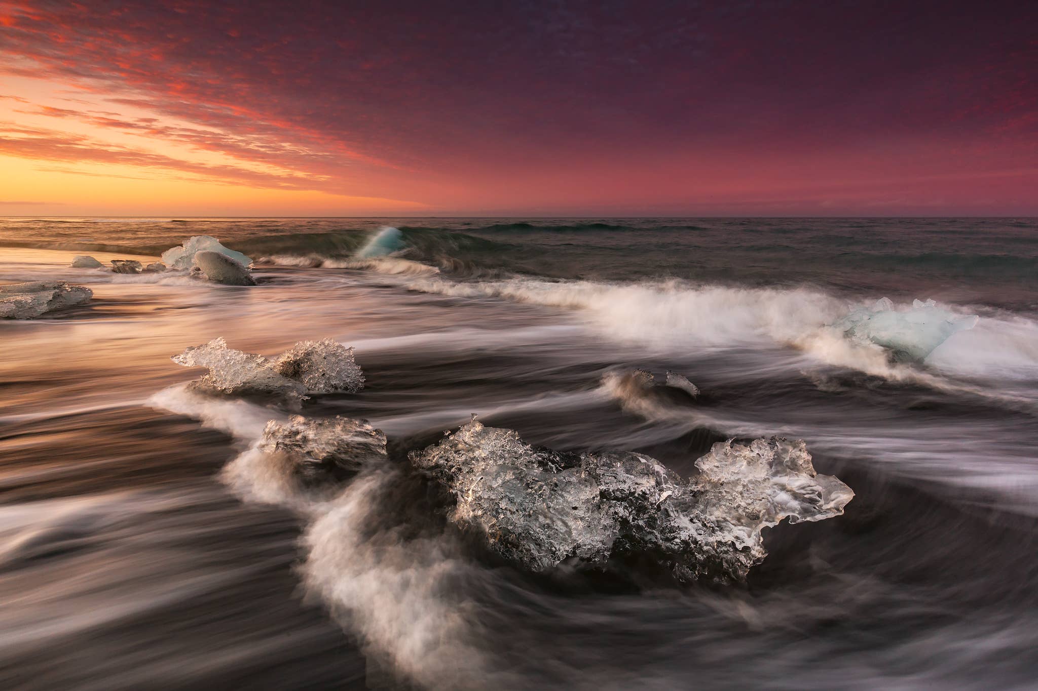 Waves swirl around glassy ice on Diamond Beach under a pink sky near Jokulsarlon Glacier Lagoon.