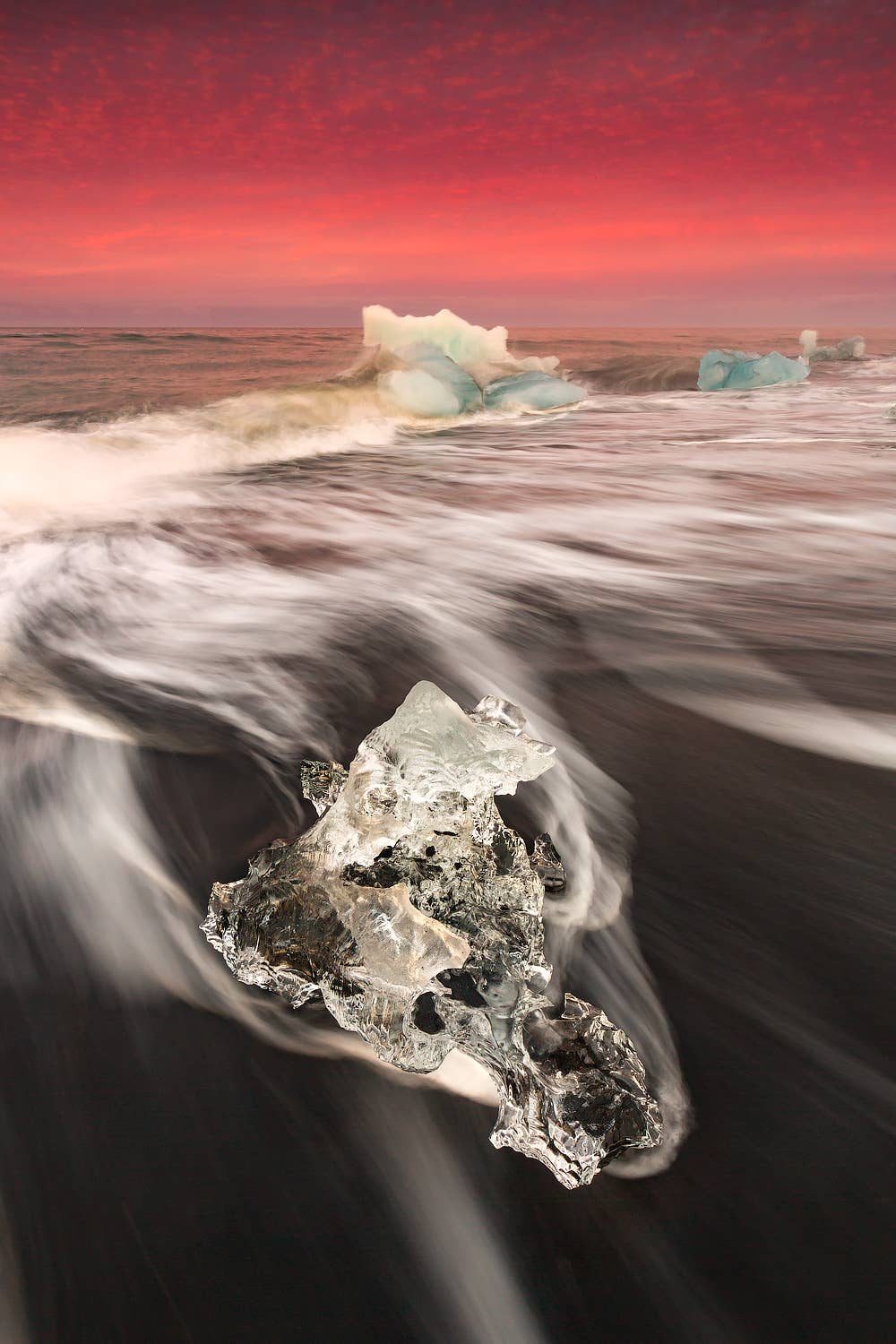 A large chunk of clear glacier ice sits in the surf of Diamond Beach under a red sky in Iceland.