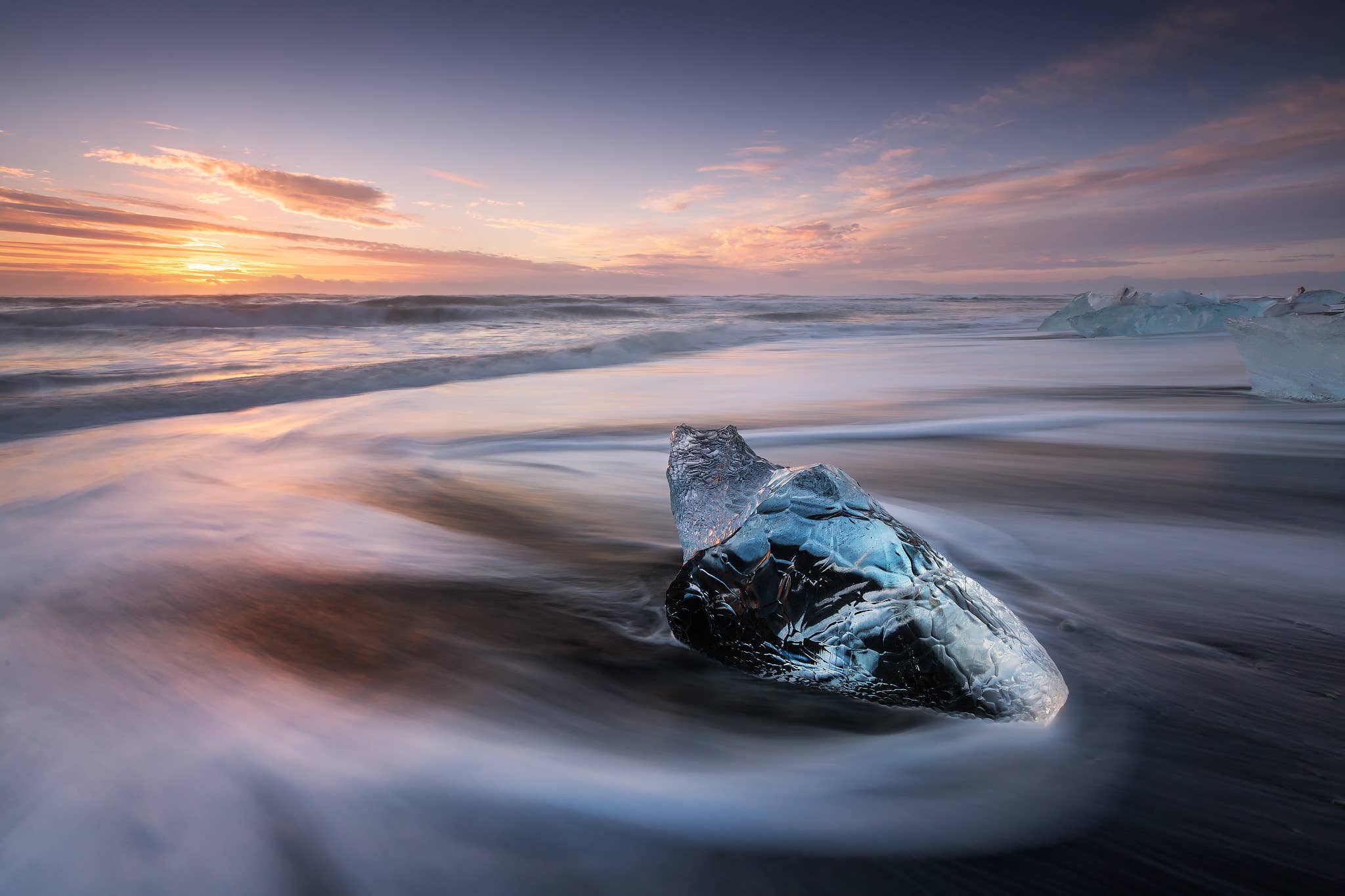 A dark piece of glacier ice glistens in the surf of Diamond Beach under a soft pastel sky in Iceland.