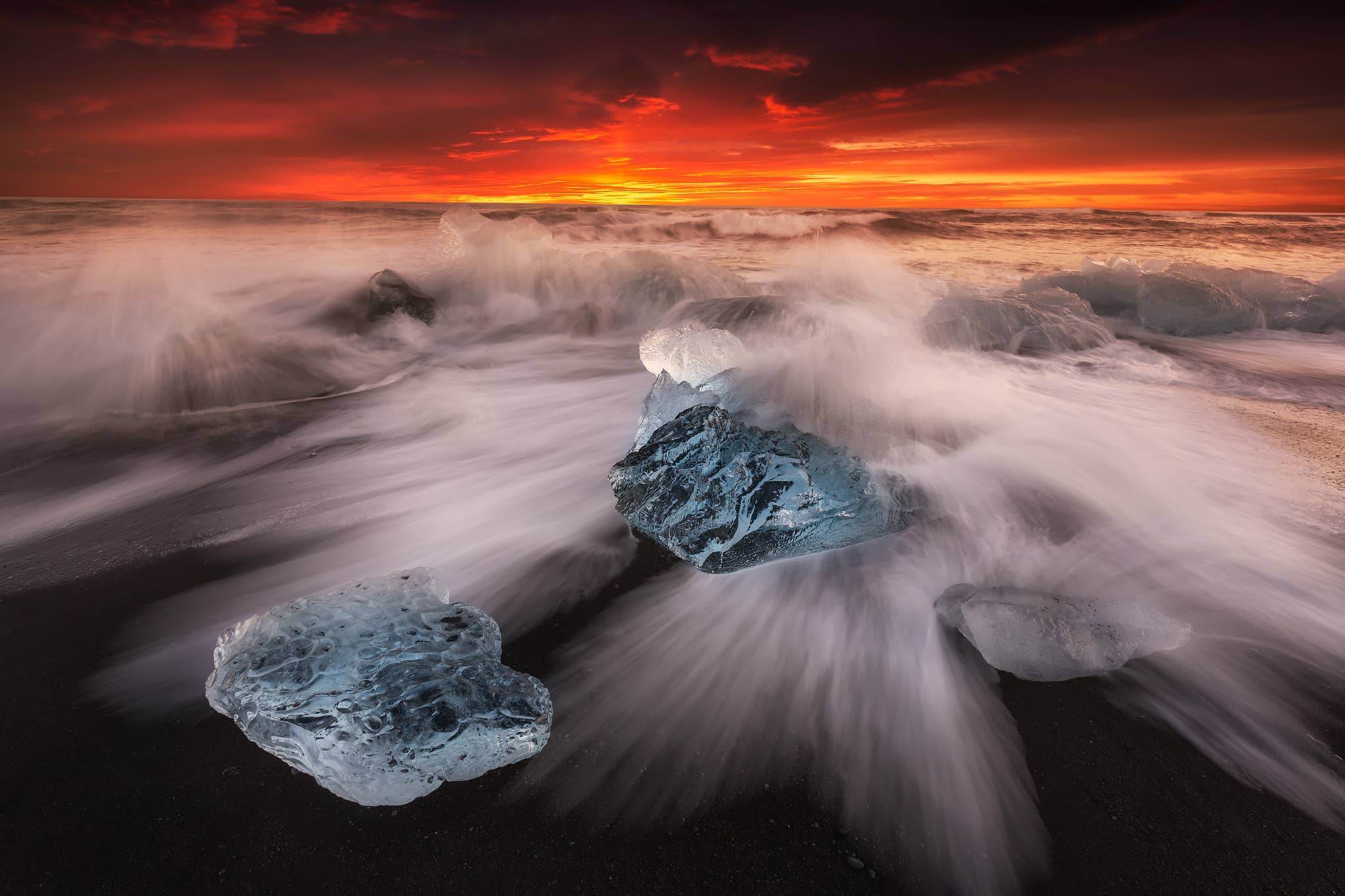 Waves rush around blue ice chunks on Diamond Beach under a fiery sky on the South Coast of Iceland.