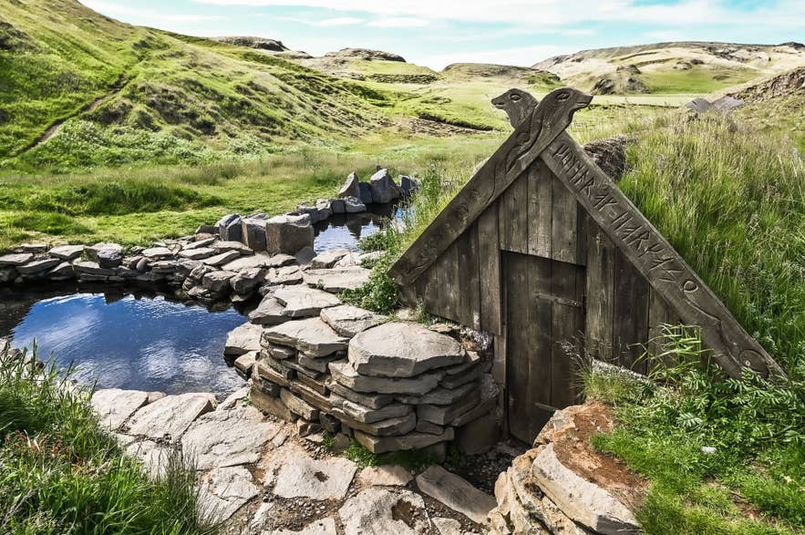 Wooden A-frame hut with a carved roof beside the stone pools at Hrunalaug Hot Spring