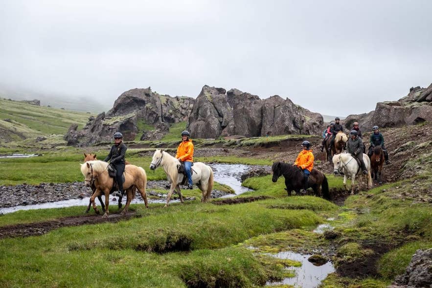 Horse riders on Eldhestar's Valley of Horses tour crossing a stream in a grassy valley in the Hengill area near Marardalur