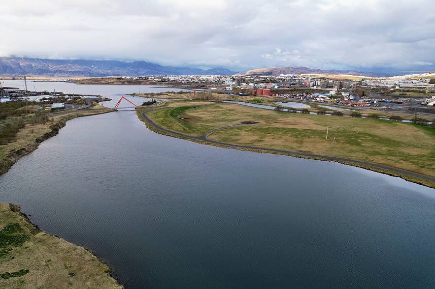 The Elliðaá river flowing past the grassy Geirsnef peninsula into Elliðaárvogur bay, with Mount Esja in the background