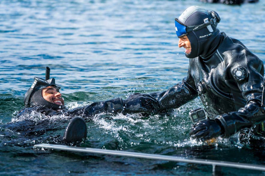 A snorkeler in a drysuit laughs in the water next to a guide at the entry platform of the Silfra fissure in Thingvellir National Park