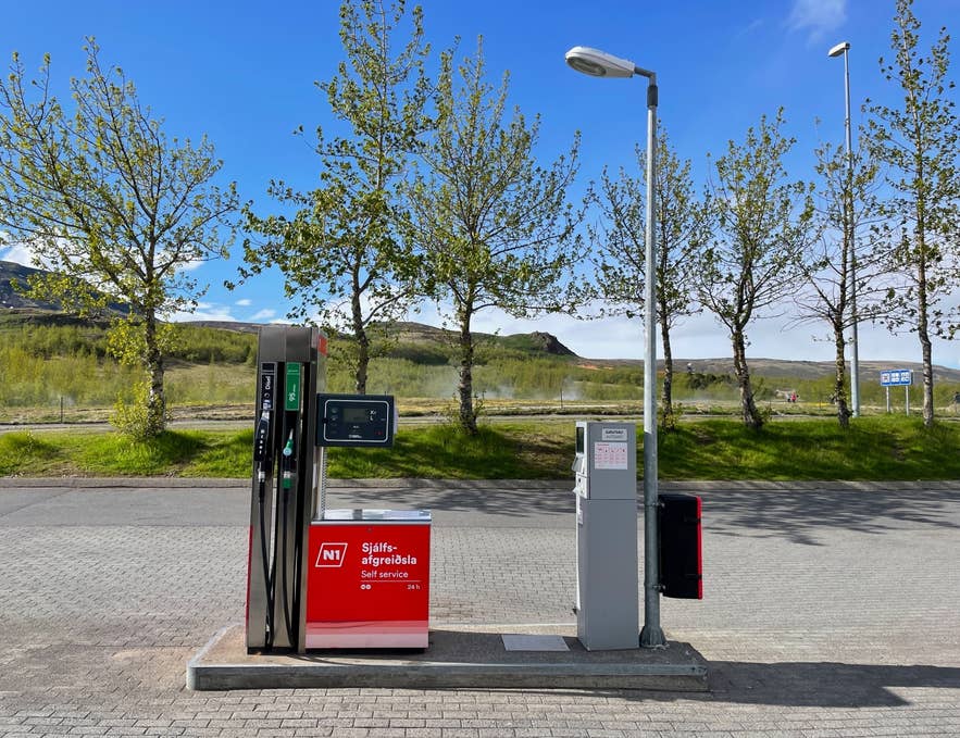 A red N1 self-service fuel pump on a paved lot with green trees and hills under a blue sky in Iceland