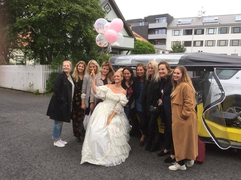 A bride stands with a bunch of balloons and big group of friends in front of a tuk-tuk in Reykjavik.