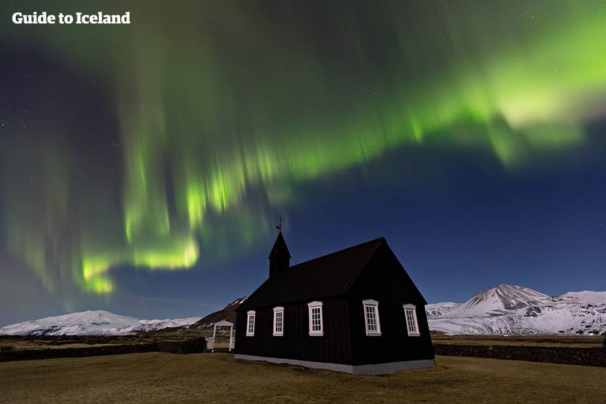 Northern lights above the black church of Budir on the Snaefellsnes Peninsula in Iceland, with snow-capped mountains in the background on a clear night.