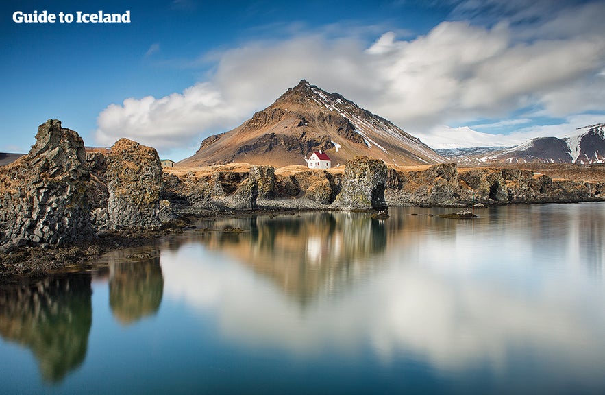 Scenic view of Arnarstapi village on the Snæfellsnes Peninsula in Iceland, featuring dramatic coastal rock formations, a lone white house, and snow-capped mountains reflected in calm waters.
