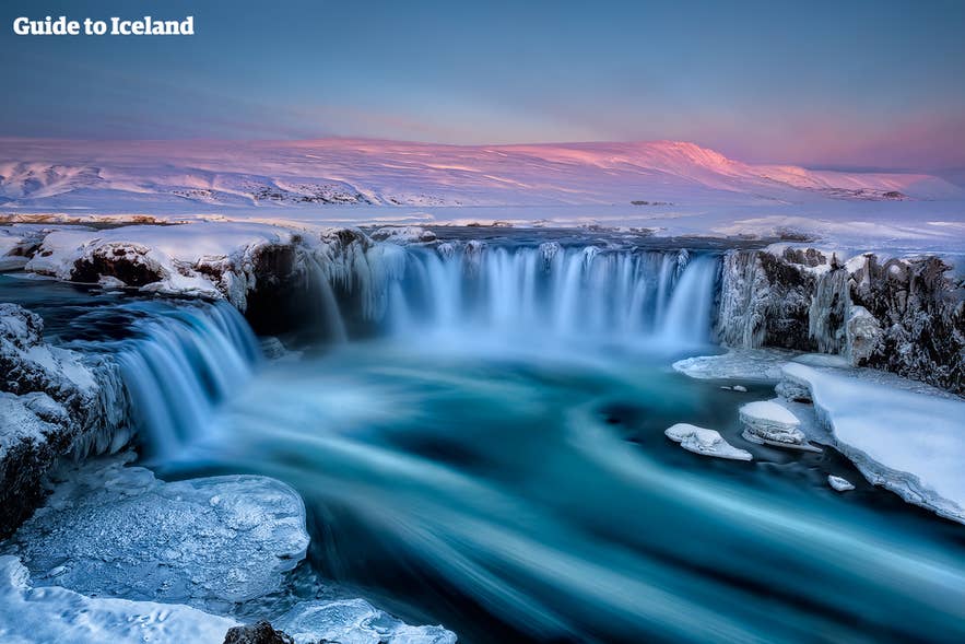 Wodospad Godafoss w północnej Islandii zimą, wśród skutej lodem scenerii, z różowym światłem wschodu słońca odbijającym się od ośnieżonych gór i turkusowej, lodowatej wody.