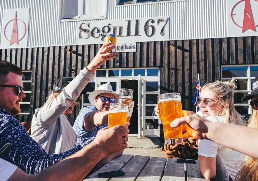 Friends toasting glasses of beer outdoors, with an Icelandic flag behind them