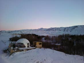 Stay warm in the cozy Eyja Mork glamping dome, evening amid snow-covered surroundings.