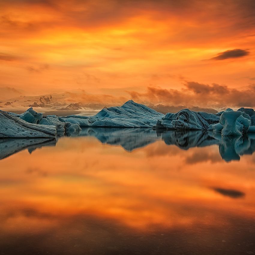 Glowing summer sunset over icebergs floating in Jokulsarlon Glacier Lagoon, Iceland, with vibrant orange skies reflecting on the still glacial waters.