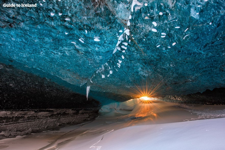 Stunning crystal ice cave in Iceland with vivid blue ice ceiling, white snow floor, and sunburst light shining through the entrance during winter.
