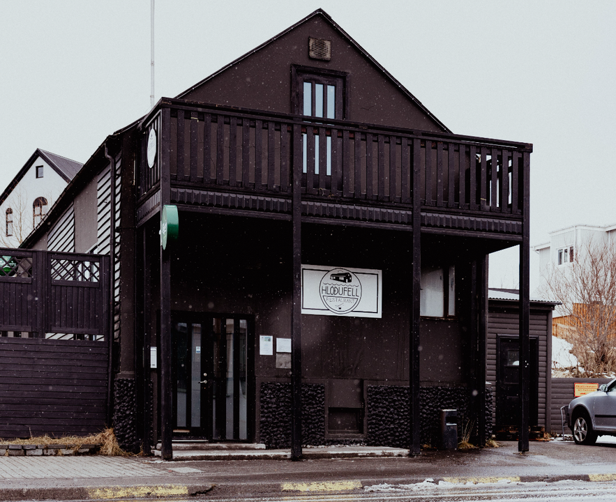 Exterior of Hlöðufell restaurant in Húsavík, Iceland, featuring a modern black timber façade with upper balcony and subtle snowfall in the scene.