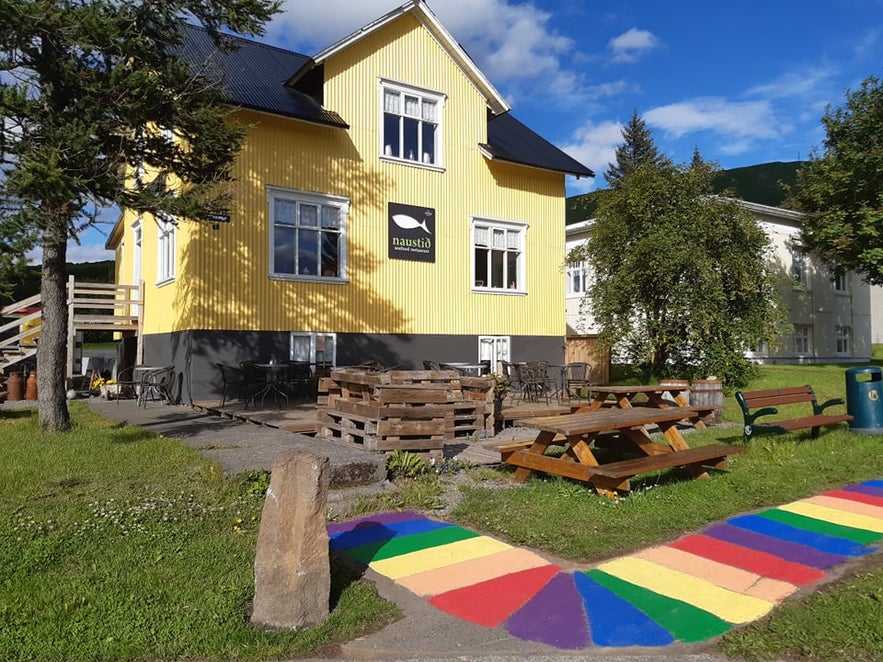 Bright yellow exterior of Naustið seafood restaurant in Húsavík, Iceland, with outdoor picnic seating and a rainbow-painted walkway in front.