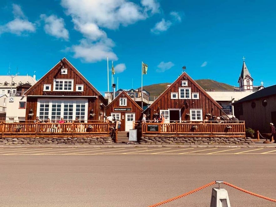 Wooden exterior of Gamli Baukur restaurant in Húsavík, Iceland, featuring traditional architecture, outdoor seating, and views of the harbor and church in the background.
