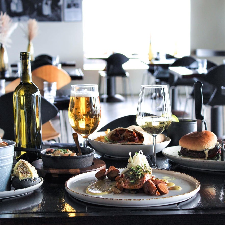 Table filled with gourmet seafood, burgers, and drinks at Sjávarborg restaurant in Hvammstangi, Iceland, with modern black-and-white interior and natural light from large windows.
