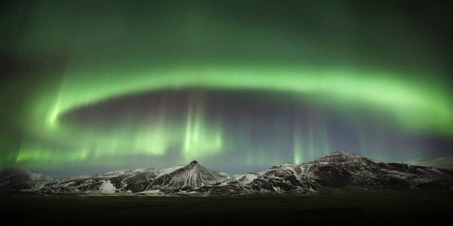 En grön norrskensbåge ovanför det snötäckta Skardsheidi-massivet i Hvalfjordur.