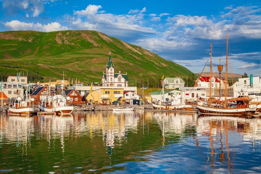 Colorful harbor in Húsavík, North Iceland, with traditional boats, charming buildings, and the iconic wooden church against a green hillside backdrop.
