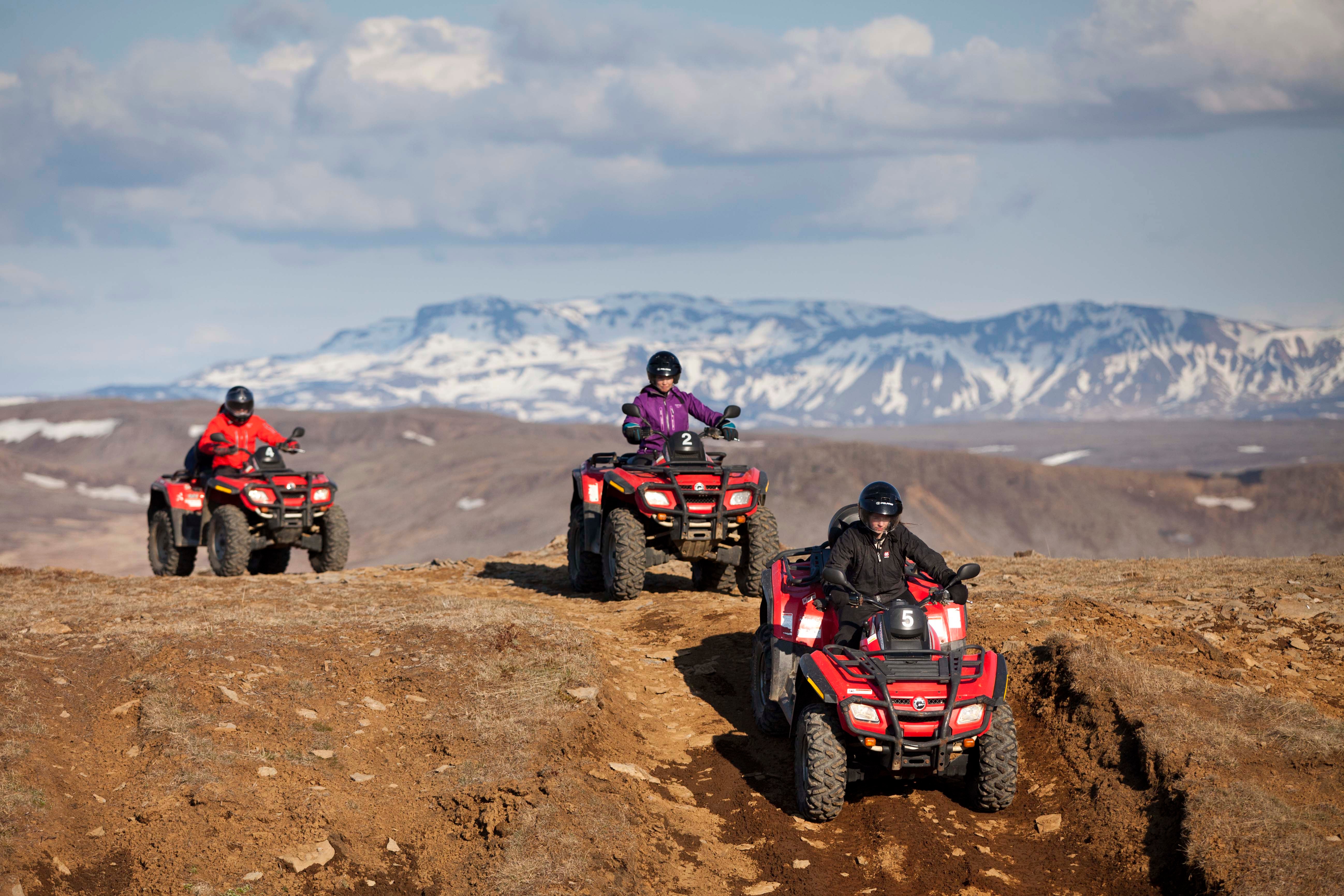 People drive ATVs over Iceland's rugged terrain with a backdrop of snowy mountains.