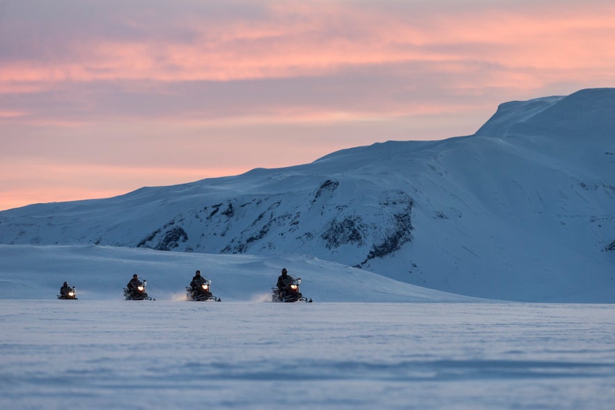 Group of travelers snowmobiling across Langjokull Glacier in West Iceland during a pink winter sunset.