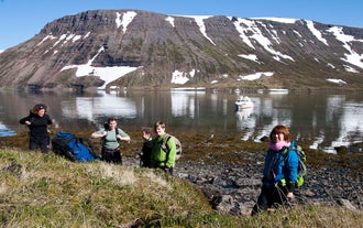 En gruppe mennesker på en tur i Hornstrandir Naturreservat står foran en fjord med en båd i.