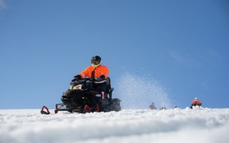 Jemand in einem orangefarbenen Overall und mit einem Helm fährt mit einem Schneemobil über den Gletscher Langjökull.