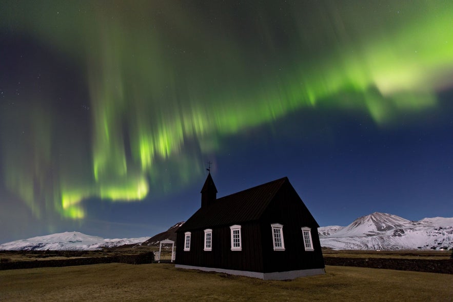 Northern lights above Budir Black Church in Snaefellsjokull National Park, with snow-covered mountains in West Iceland.